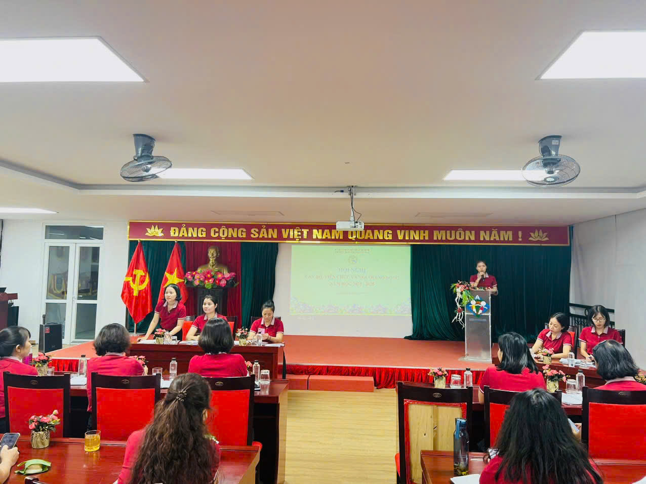 A group of people sitting at a podium in a room with a stage and a screenDescription automatically generated